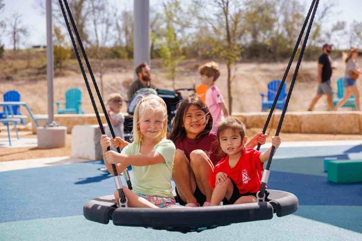Three children sitting on a round rope swing at a playground, with adults and other children in the background.