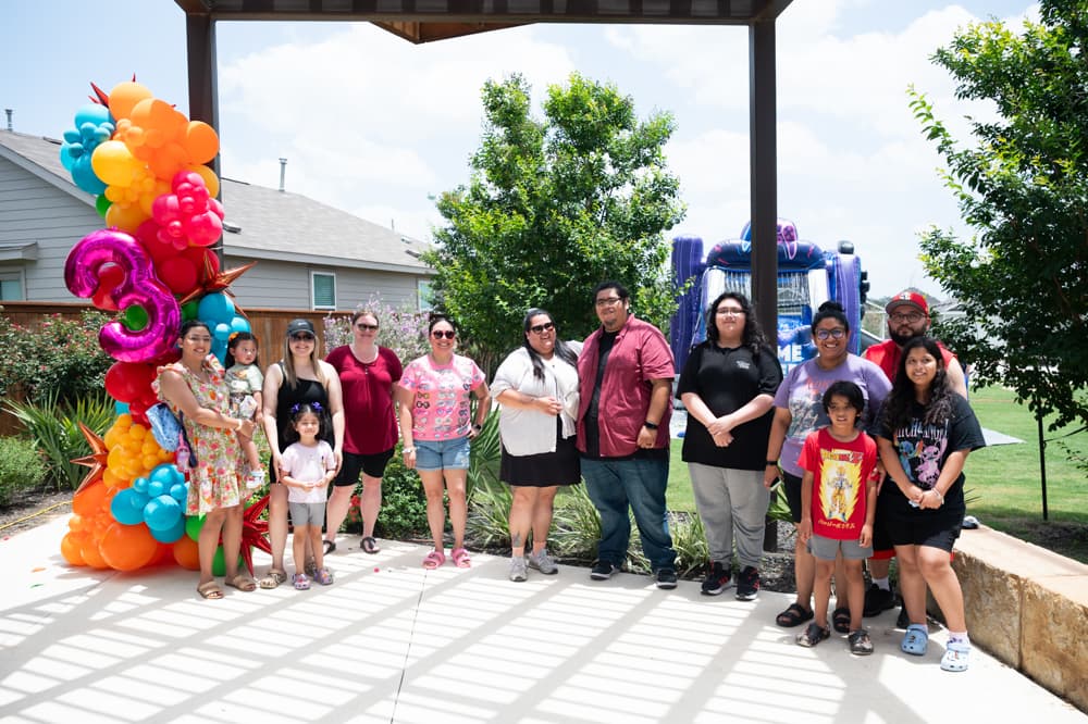 A group of people pose for a photo outdoors under a pergola at the VIDA San Antonio development, with balloons shaped as the number three and a bounce house visible in the background.