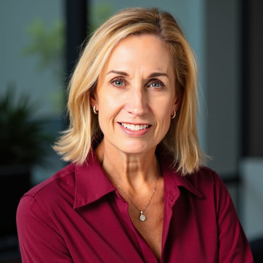 A woman with shoulder-length blonde hair, wearing a maroon blouse and a pendant necklace, smiles at the camera in an indoor setting.